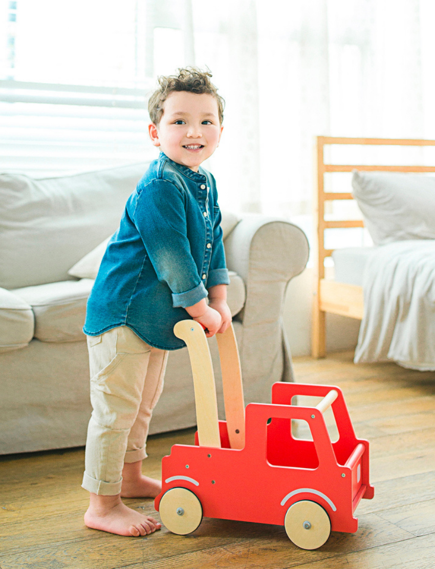 The Moover Push Truck is a red wooden toy car featuring beige wheels and handlebars. It boasts a simplistic design with cut-out windows and a small basket area at the back, marked with the word "LINE." This toy is designed for children to push or pull.