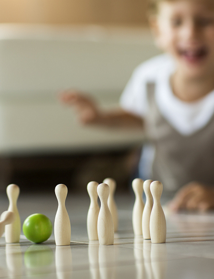 Image of a small, wooden bowling set perfect for tabletop ten-pin bowling. The Milaniwood "Green Mini Bowling" set includes ten light wooden pins arranged in a triangular formation, with two wooden balls—one green and the other light wood—in the foreground along with a small round wooden base. Ideal for family game nights.