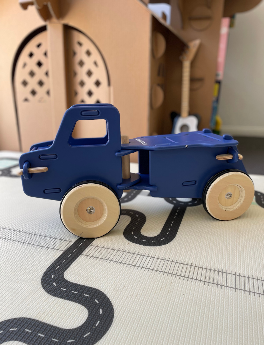 The Moover Classic Dump Truck is a light blue wooden vehicle featuring four sturdy wooden wheels. Its front is equipped with round headlights and a driver's cab, while the rear boasts a spacious flatbed for carrying items. This durable toy encourages fine motor skills and is perfect for young children.