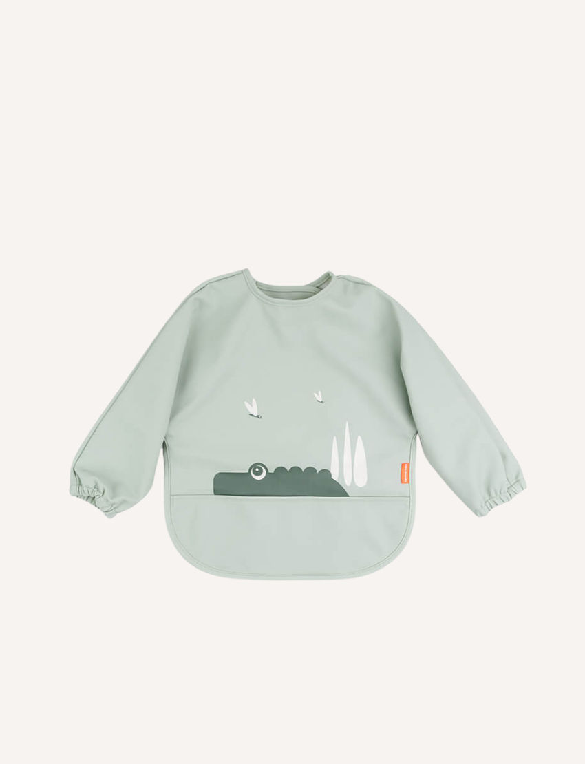 The Done by Deer Sleeved Pocket Bib is waterproof and features a light green, long-sleeve design with alligators, trees, and birds. It has a curved bottom edge, smooth fabric, gathered cuffs, and a small right-side tag—perfect for easy-to-clean mealtime fun.