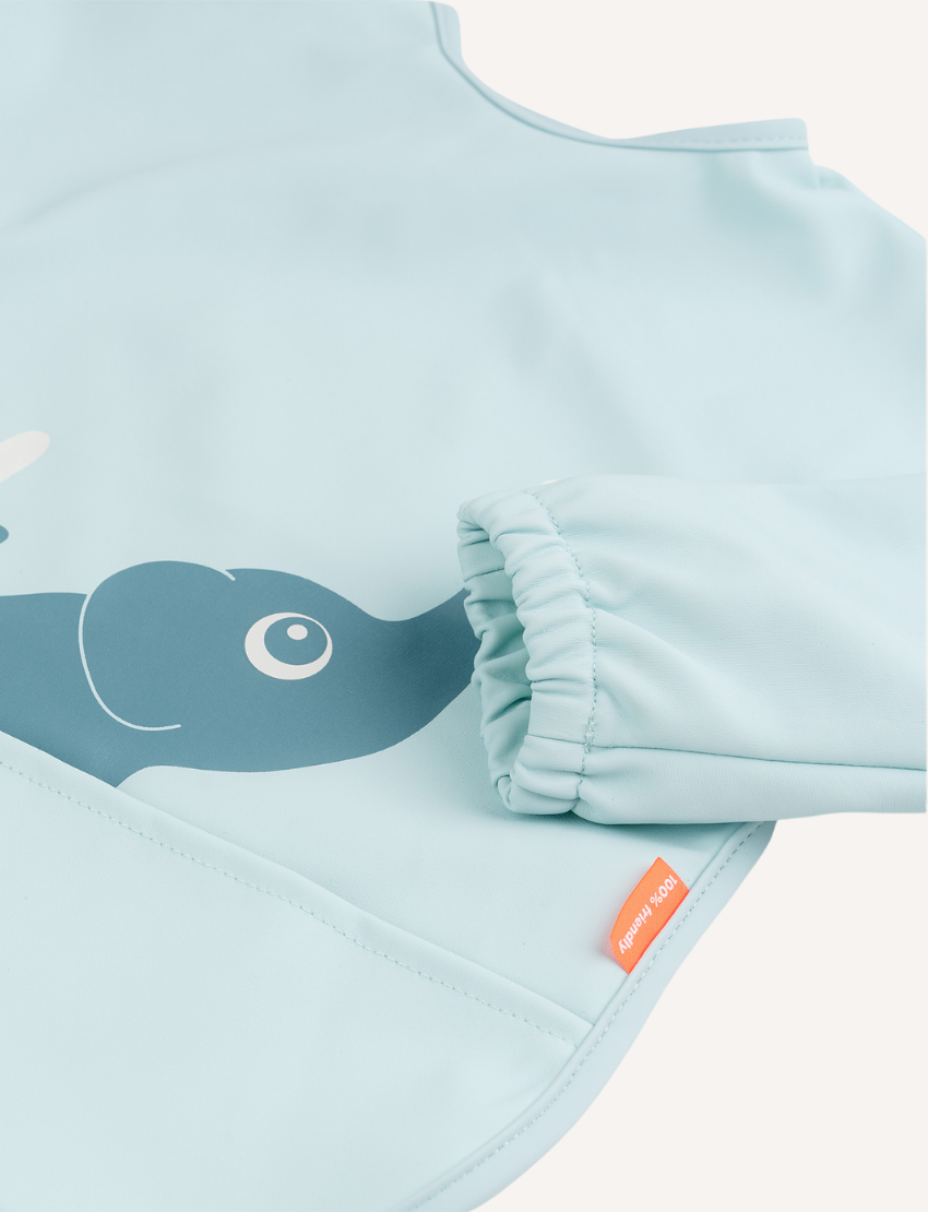 Done by Deer presents the Sleeved Pocket Bib in green, showcasing a minimalist crocodile and bird design. It is waterproof with elastic cuffs and features an adjustable closure, a bottom pocket, and a small orange tag on the right side.