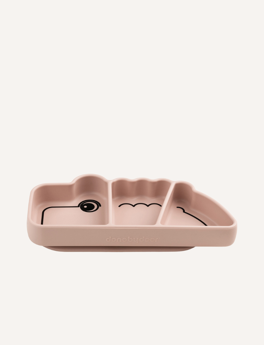 A pink plastic children's plate designed to look like a crocodile, with the plate divided into three sections, one of which includes the crocodile's eye and another its foot. The brand name "Done by Deer" is embossed on the front. The product name is "Stick & Stay Plate - Crocodile.