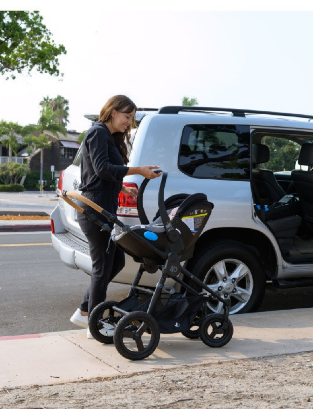 The Bumbleride ERA Capsule Adaptors are displayed in a profile view. The silver and black stroller, equipped with four wheels, features a Maxi-Cosi car seat attached to it and a wooden handlebar. The bottom of the stroller has a visible mesh storage basket, and the black wheels are fitted with rubber tires.
