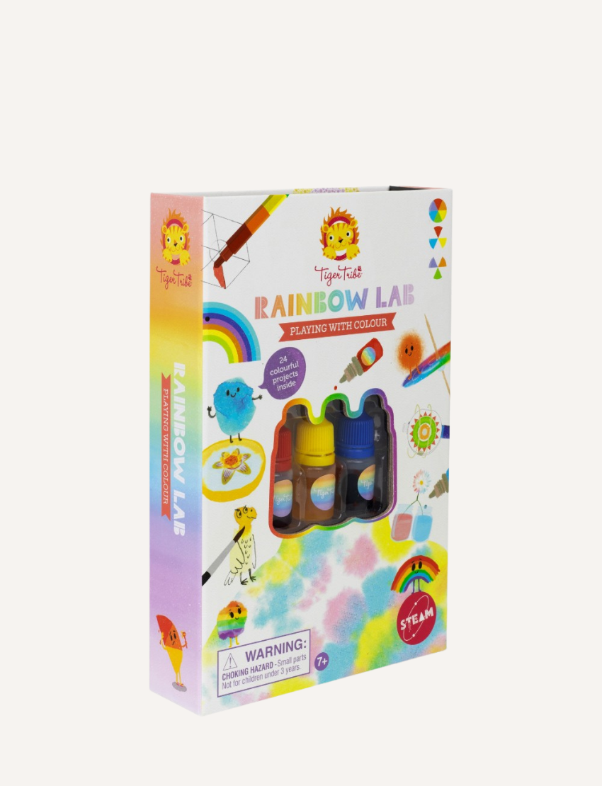 A DIY craft kit from Tiger Tribe is displayed, including a Rainbow Lab - Playing with Colour booklet titled "DIY Thaumatrope" with a page open to project instructions. Surrounding items include markers, paint bottles, a brush, and papers featuring rainbow and cloud designs.