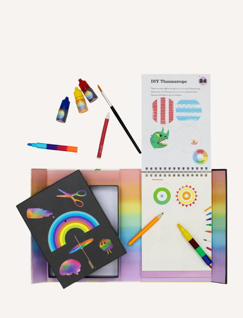 A DIY craft kit from Tiger Tribe is displayed, including a Rainbow Lab - Playing with Colour booklet titled "DIY Thaumatrope" with a page open to project instructions. Surrounding items include markers, paint bottles, a brush, and papers featuring rainbow and cloud designs.