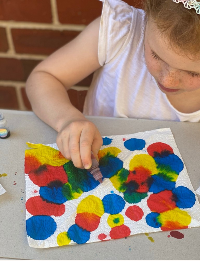 A DIY craft kit from Tiger Tribe is displayed, including a Rainbow Lab - Playing with Colour booklet titled "DIY Thaumatrope" with a page open to project instructions. Surrounding items include markers, paint bottles, a brush, and papers featuring rainbow and cloud designs.