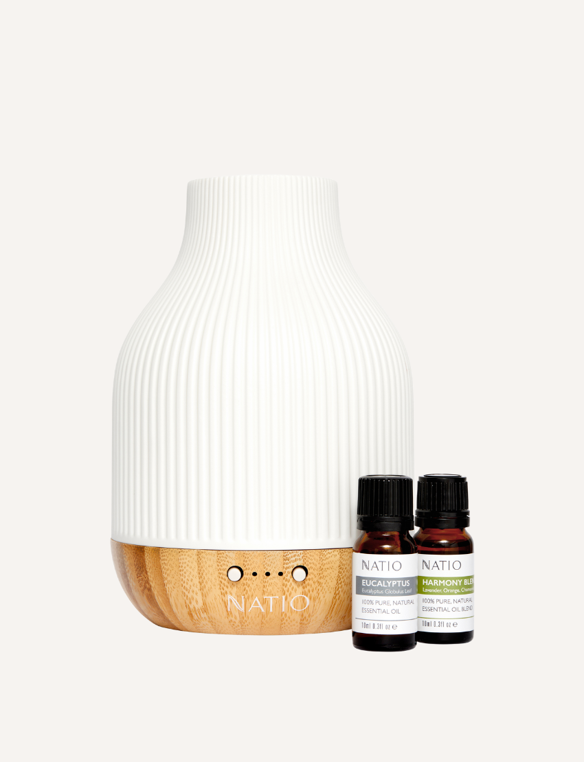 A white, ribbed Harmony Essential Oil Diffuser Gift Set from Natio, featuring a wooden base, is displayed next to two small bottles of essential oils labeled "Natio Eucalyptus" and "Natio Harmony" on a plain, neutral background. It promises a serene aromatherapy experience through advanced ultrasonic technology.