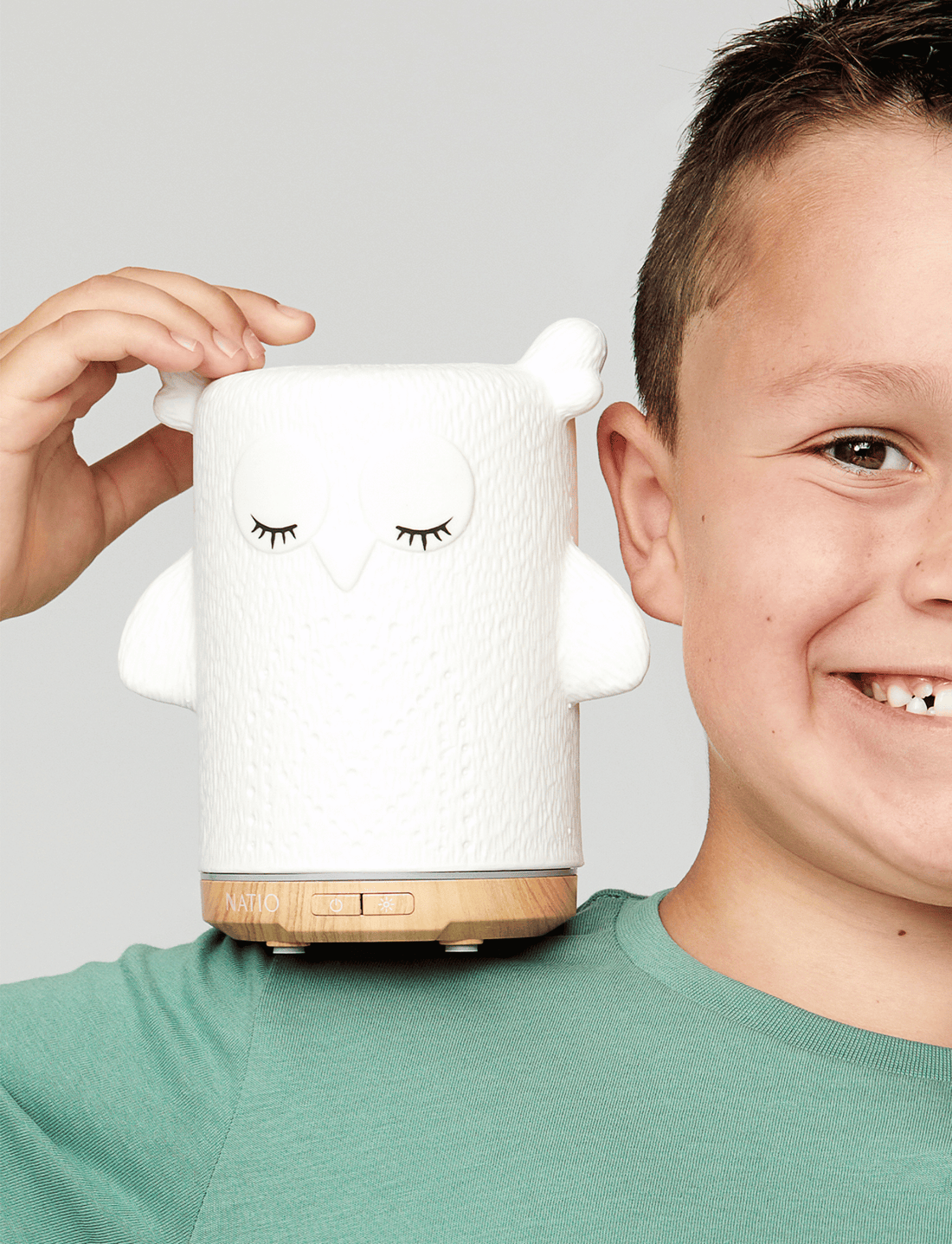 A white ceramic owl-shaped essential oil diffuser, ideal for children's aromatherapy, is placed next to its box. The box showcases "Natio Kids" along with an image and description of the diffuser. With its woodgrain-look base and charming owl design, the Kids Olive the Owl Ultrasonic Diffuser from Natio is perfect for any child's room.