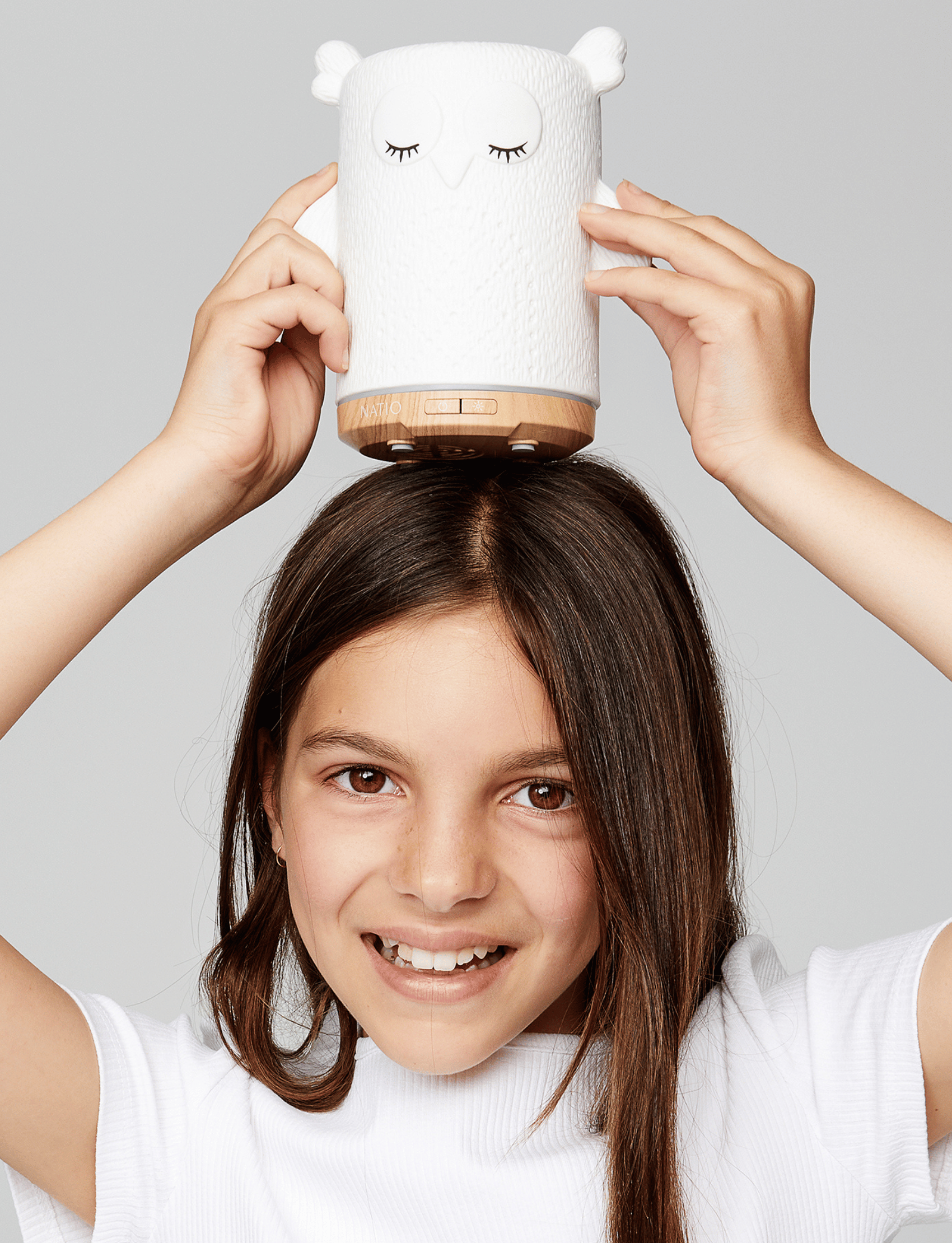 A white ceramic owl-shaped essential oil diffuser, ideal for children's aromatherapy, is placed next to its box. The box showcases "Natio Kids" along with an image and description of the diffuser. With its woodgrain-look base and charming owl design, the Kids Olive the Owl Ultrasonic Diffuser from Natio is perfect for any child's room.