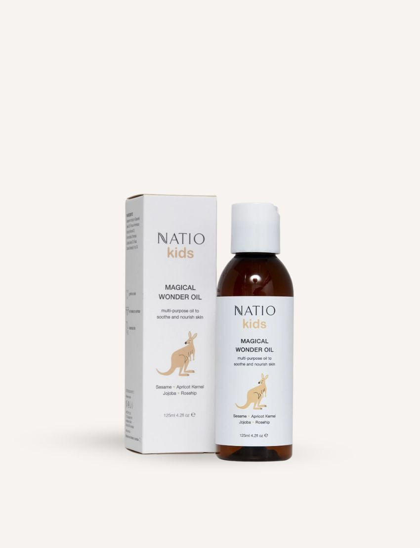 A white bottle of Natio Kids Magical Wonder Oil is positioned beside its box. The packaging showcases a kangaroo illustration and text that highlights the inclusion of sesame, apricot kernel, jojoba, and rosehip oils. It promises nourishing skin benefits and aromatherapy for kids. The bottle has a capacity of 125ml (4.2 fl oz).