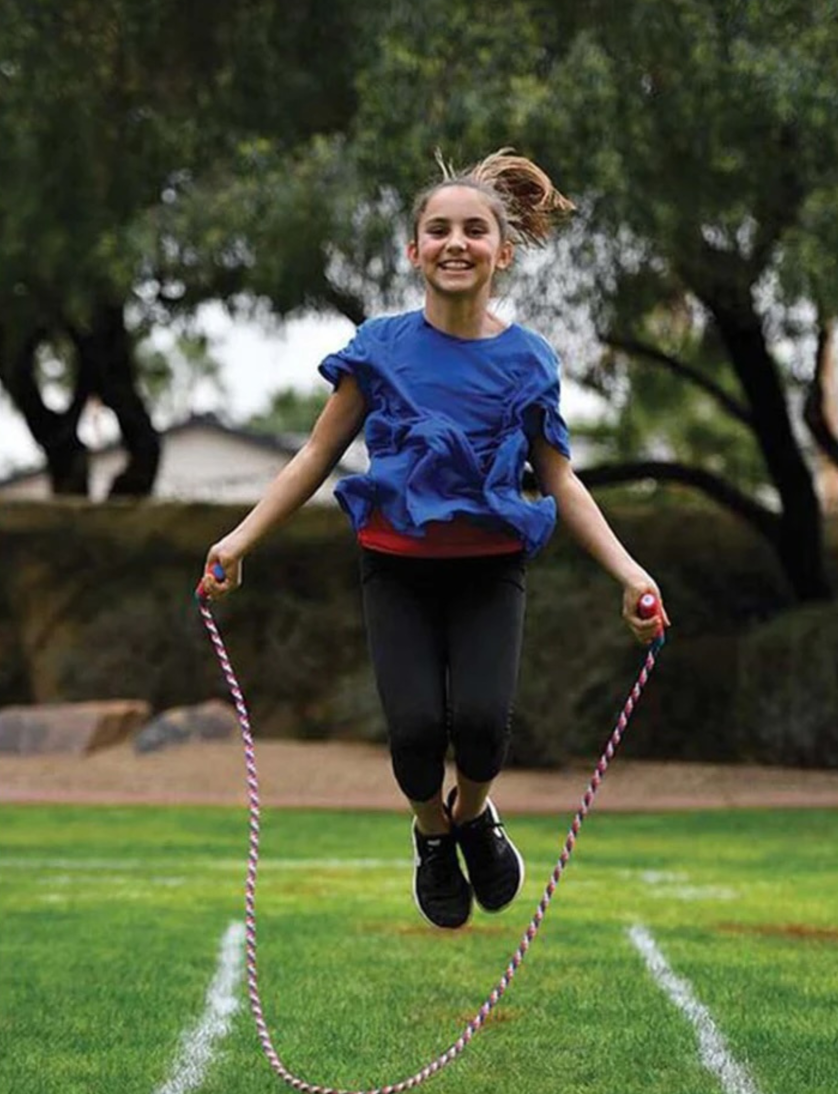 A girl with face paint, wearing a red top and black leggings, crawls under a series of red string lines set at various heights in an outdoor grassy area. She is smiling and appears to be participating in Slackers' Ninja Obstacle Course with Bounce Balls, perfect for some family fun.