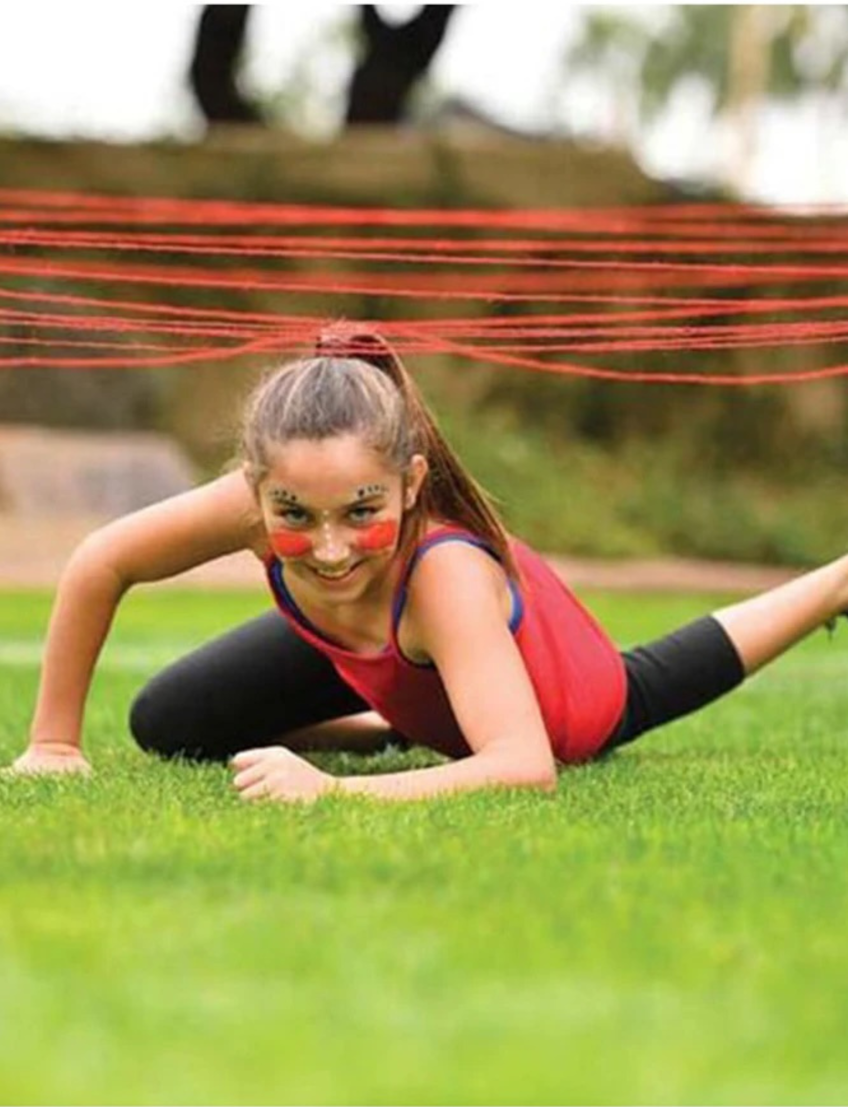 A girl with face paint, wearing a red top and black leggings, crawls under a series of red string lines set at various heights in an outdoor grassy area. She is smiling and appears to be participating in Slackers' Ninja Obstacle Course with Bounce Balls, perfect for some family fun.