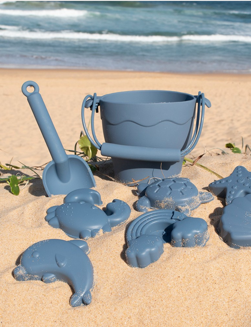 The Playground 8-Piece Bucket & Spade Set features a BPA-free silicone brown bucket, spade, five animal molds (cloud, rainbow, crab, shell, turtle), and a starfish mold. A white drawstring bag is included for easy storage—perfect for sandcastle crafting!.