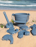 Playground's 8-Piece Bucket & Spade Set, featuring a yellow bucket with a handle, a small shovel, five sand molds in various shapes (sun, cloud, starfish, turtle, and crab), and a drawstring storage bag—all neatly arranged on a plain background.