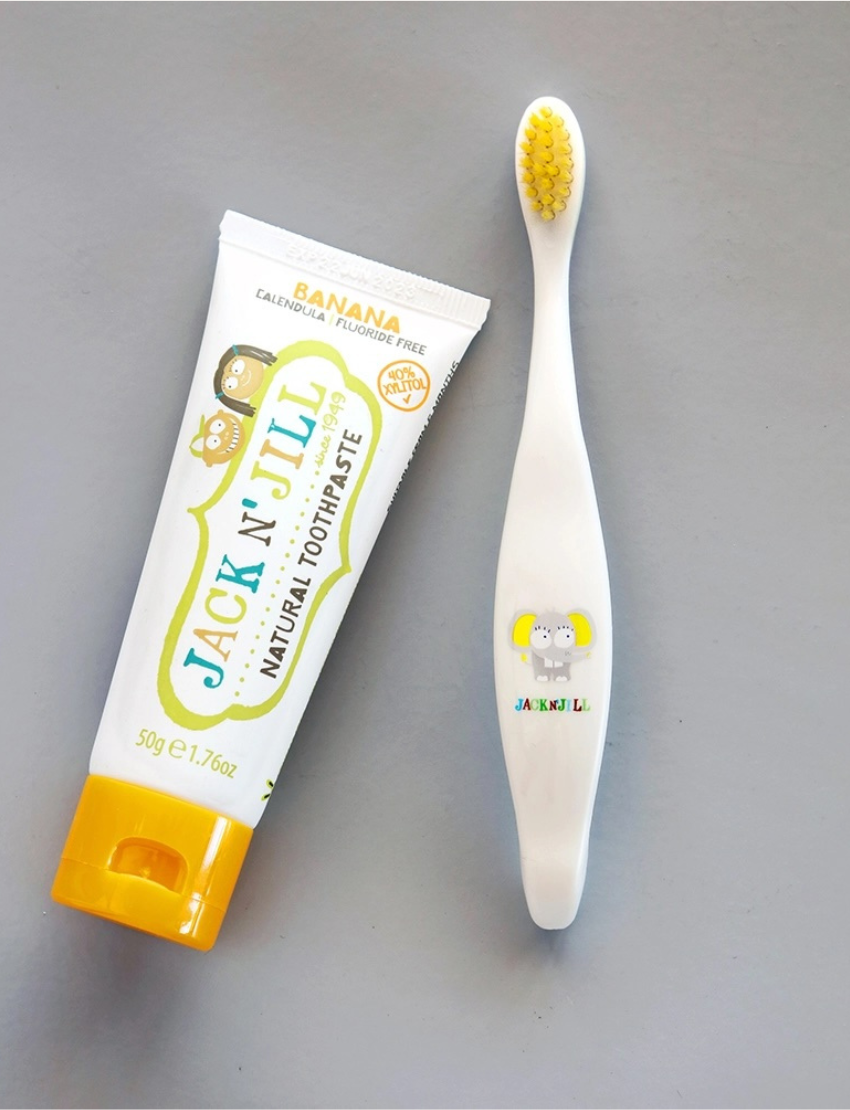 A Jack N' Jill Biodegradable Toothbrush, designed for children with a koala theme, and a tube of Jack N' Jill natural banana-flavored toothpaste adorned with cartoon animals on the label, are laid out on a gray surface.