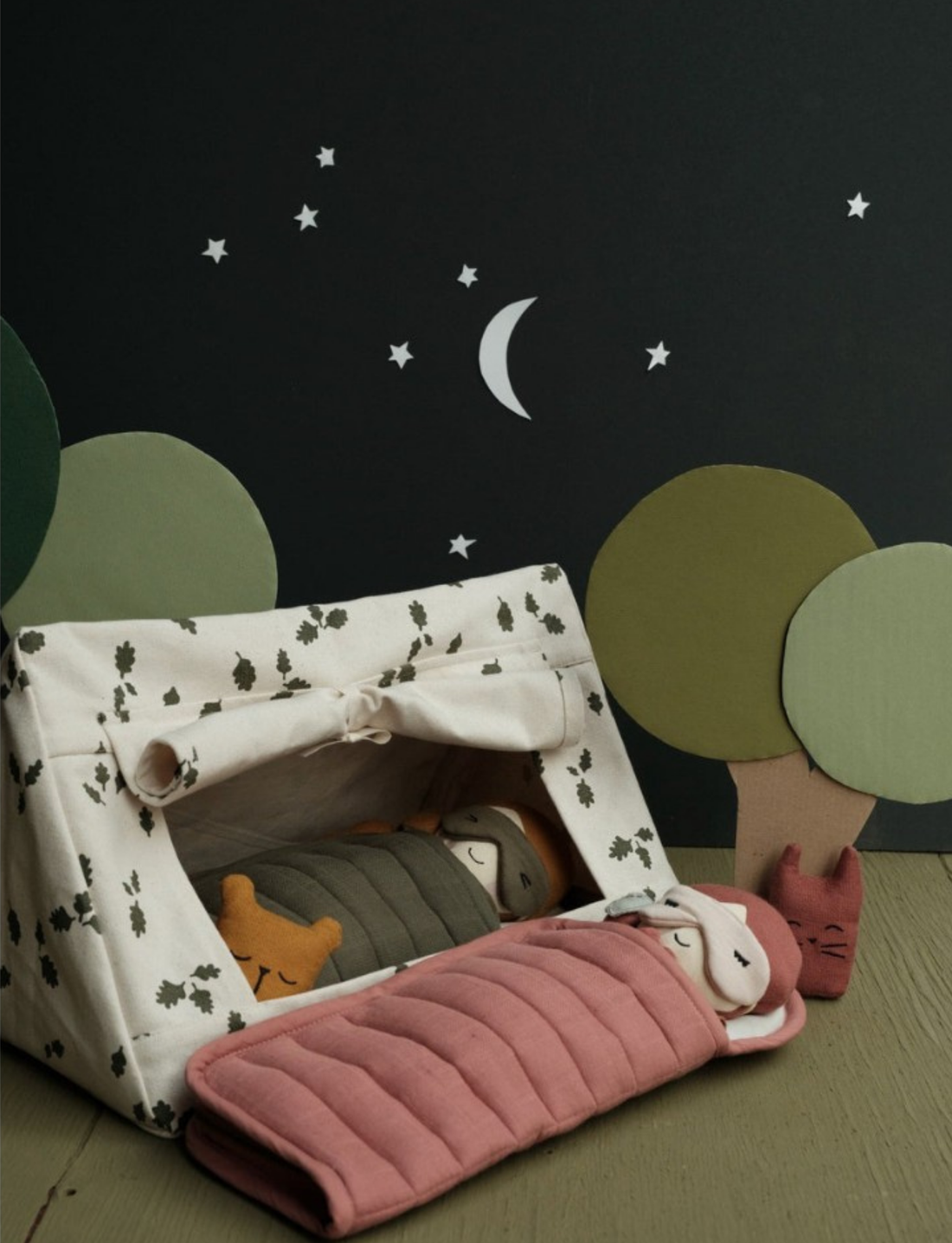 Inside the Doll Tent - Oak Leaves by Fabelab, two soft dolls lie in sleeping bags—one green and one pink. The tent, made of organic cotton canvas, is adorned with leaf patterns. The backdrop showcases a crescent moon and stars against a black sky, with paper trees on either side, creating a serene nighttime camping scene.