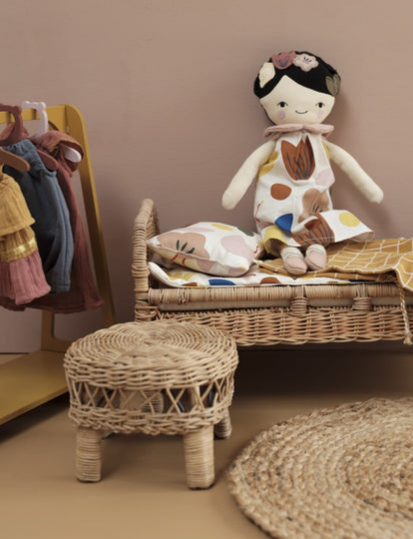 A hand, clad in an orange sweater, holds a Fab Friends Doll from the brand Fabelab. The fabric doll features dark brown skin and curly hair styled in two pigtails. She is dressed in a yellow outfit decorated with a sun design and wears matching yellow shoes. The background is a light brown wall.