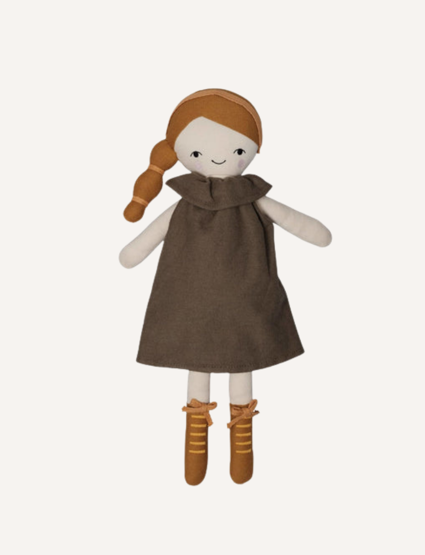 A hand, clad in an orange sweater, holds a Fab Friends Doll from the brand Fabelab. The fabric doll features dark brown skin and curly hair styled in two pigtails. She is dressed in a yellow outfit decorated with a sun design and wears matching yellow shoes. The background is a light brown wall.