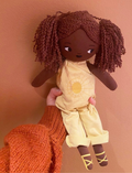 A hand, clad in an orange sweater, holds a Fab Friends Doll from the brand Fabelab. The fabric doll features dark brown skin and curly hair styled in two pigtails. She is dressed in a yellow outfit decorated with a sun design and wears matching yellow shoes. The background is a light brown wall.