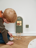 The Christmas Elf Door - Green with Light by Fabelab features a small, green door with a gold handle. Accompanied by a tiny ladder and a matching green mailbox adorned with a white decorative emblem, the door boasts four window panes and is decorated with a bow and floral design at the top, making it ideal for welcoming Christmas elves.