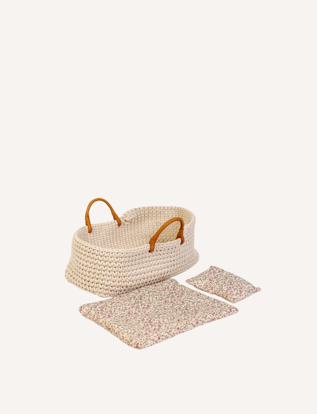 The Doll Knitted Basket and Bedding Set by Astrup features a woven, cream-colored baby Moses basket with two light brown handles. Meticulously crafted from 100% cotton, it comes with a matching floral blanket and pillow made from mille fleur fabric, neatly arranged outside the basket. The items are displayed against a plain white background.