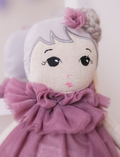 The Doll Fleur and Change of Outfit by Astrup features a soft fabric doll with light skin, embroidered eyes, short white hair adorned with a small flower, dressed in a purple tulle outfit. To the left of the doll lies a separate pink lace and tulle dress displayed on a flat surface.