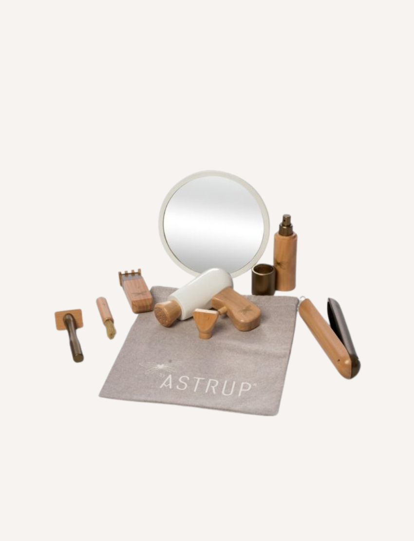 The Wooden Role Play Doll Hairdressing Set by Astrup includes a handheld mirror, a wooden razor, a brush, a comb, scissors, a spray bottle, a trimmer, and various small accessories. These tools are displayed on a fabric mat featuring the brand name "by Astrup.