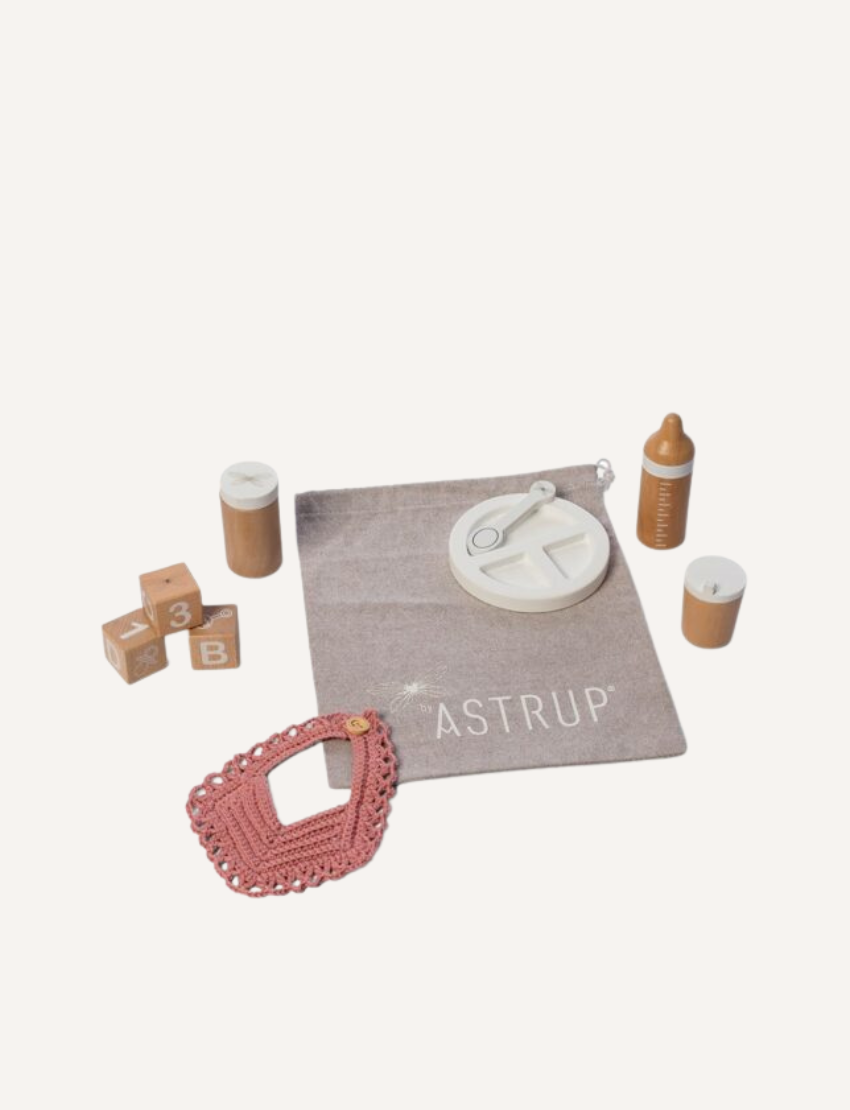 The Wooden Role Play Doll Feeding Set by Astrup features an assortment of baby items, such as lettered wooden blocks, a wooden baby bottle, wooden jars, a pink crocheted bib, and a white circular toy with a spinning mechanism. These items are displayed on a white background.