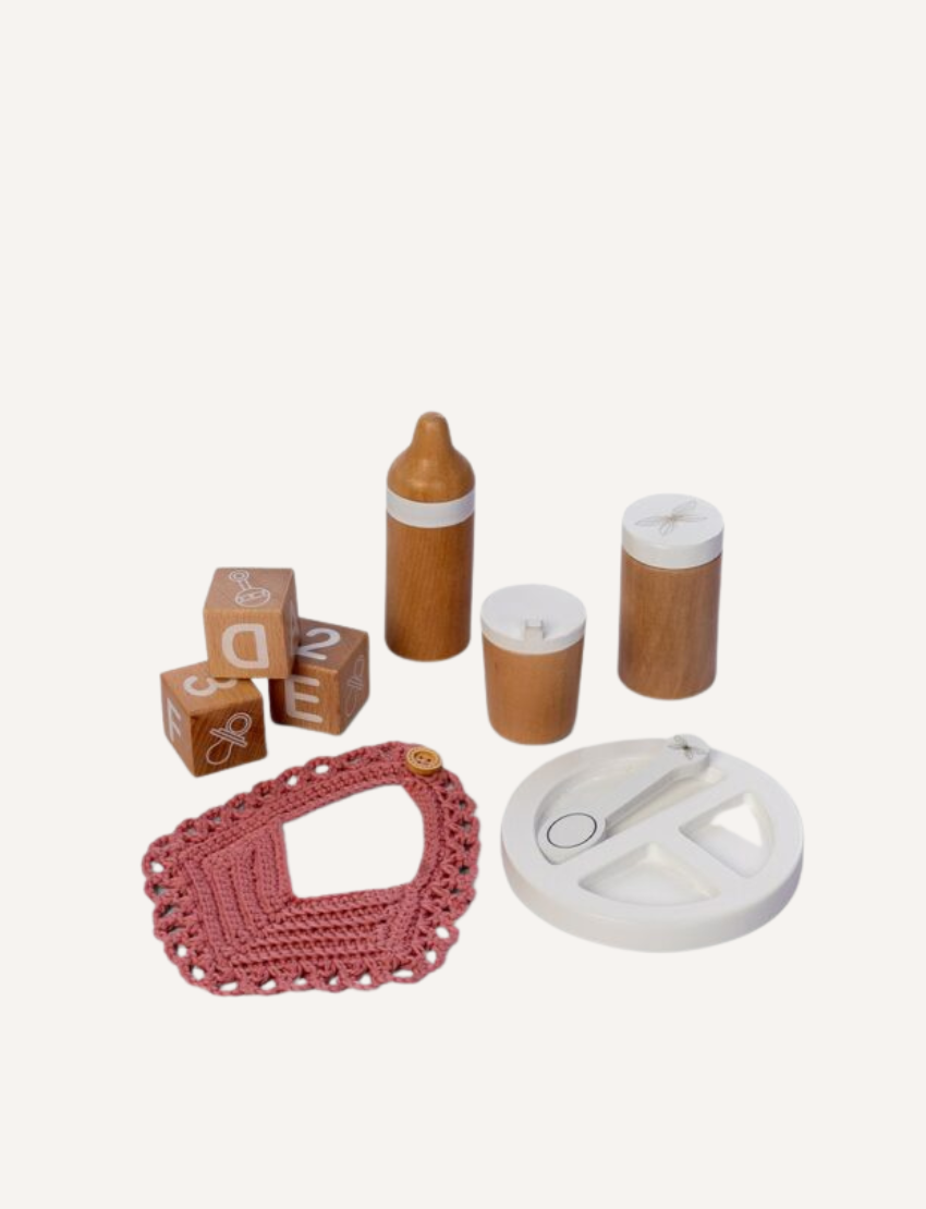 The Wooden Role Play Doll Feeding Set by Astrup features an assortment of baby items, such as lettered wooden blocks, a wooden baby bottle, wooden jars, a pink crocheted bib, and a white circular toy with a spinning mechanism. These items are displayed on a white background.