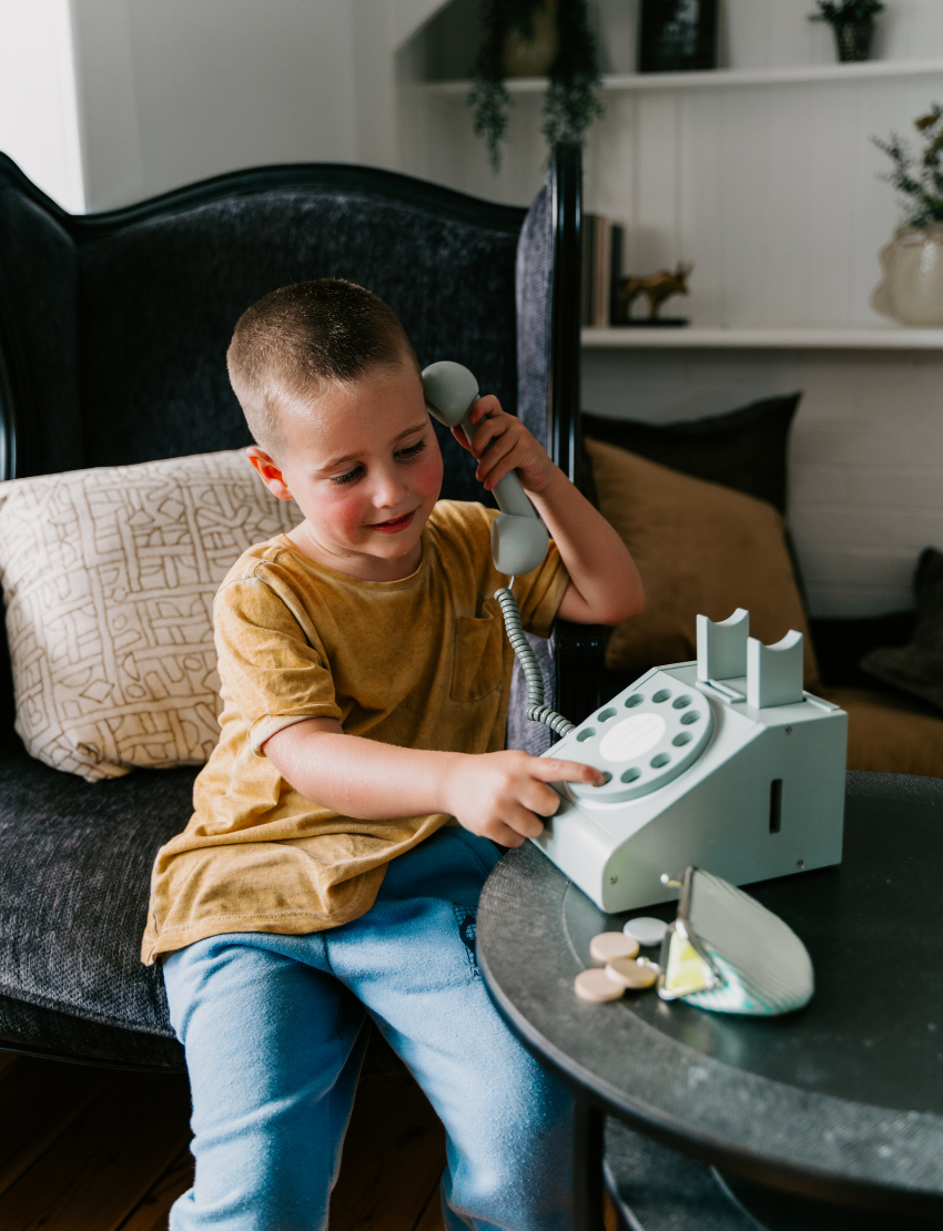 A young boy in a yellow shirt and blue pants sits on a dark armchair while holding the receiver of a kiko+ and gg* Telephone to his ear. He is dialing a number with his other hand. A small, open coin purse and some coins are on the table next to him.