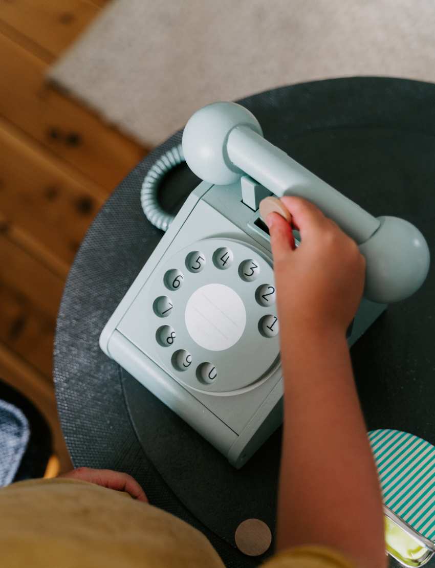 A young boy in a yellow shirt and blue pants sits on a dark armchair while holding the receiver of a kiko+ and gg* Telephone to his ear. He is dialing a number with his other hand. A small, open coin purse and some coins are on the table next to him.