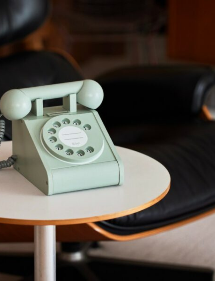 A young boy in a yellow shirt and blue pants sits on a dark armchair while holding the receiver of a kiko+ and gg* Telephone to his ear. He is dialing a number with his other hand. A small, open coin purse and some coins are on the table next to him.