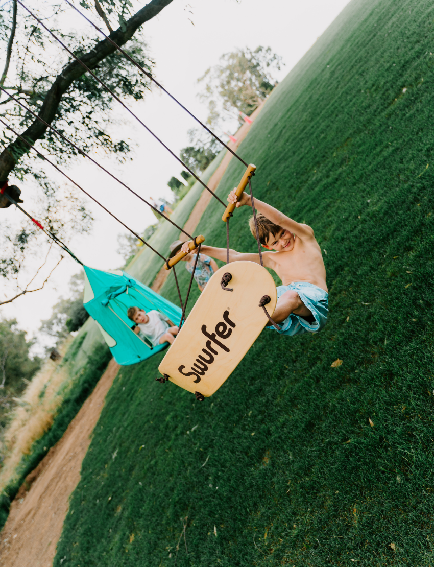 A Swurfer Swingboard by Flybar, featuring a wooden curved board with the brand name "Flybar" printed on it. It comes with two sturdy white ropes accented in orange and two wooden handles. The board includes four holes for rope attachment.