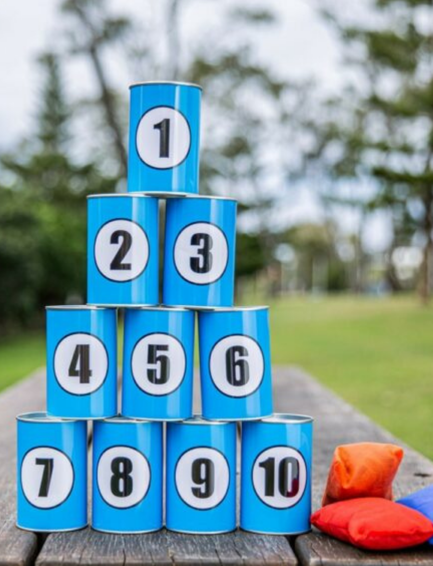 A pyramid of Throwing Cans by Formula Sports and Games, numbered 1 to 10, sits on a wooden table outdoors. Two bean bags, one red and one orange, are placed next to the cans. Trees and grass are visible in the background.