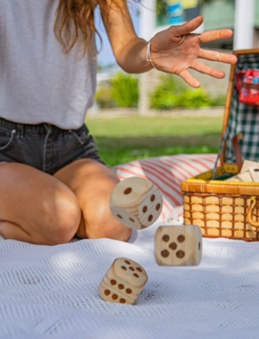 A cylindrical container labeled "Outdoor Yatzy" by Formula Sports and Games, featuring a blue and black design. It states: "5 dice, each measuring 5 x 5 x 5 cm, made from high-quality wood, with a score sheet included." A photo of someone playing dice on the grass is visible on the lower section.