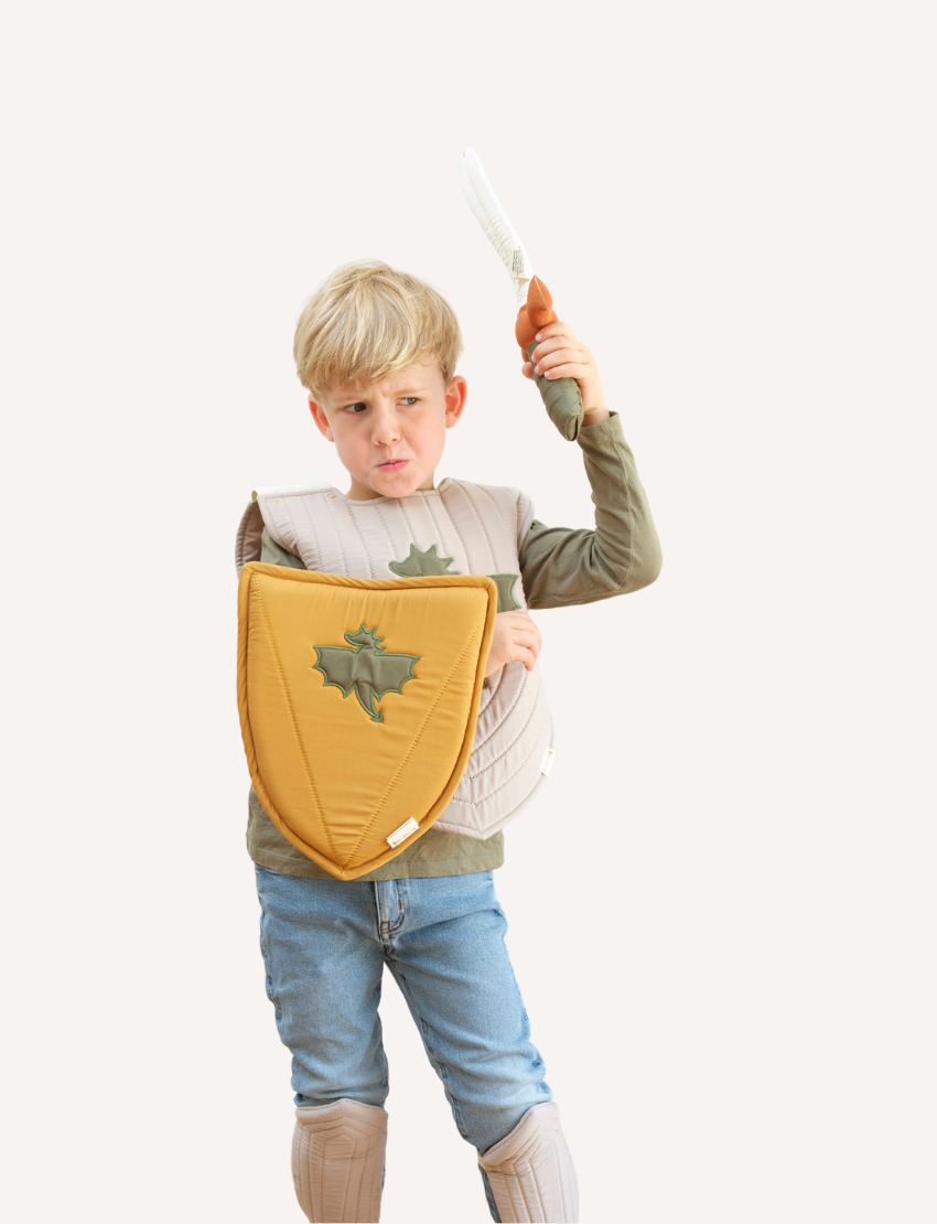 A young boy wears medieval-style costume armor, complete with a light brown chest piece and matching shin guards. He holds the yellow Shield & Sword by Fabelab in one hand, featuring a green leaf emblem, and poses as if mid-action.