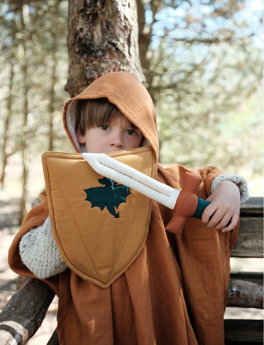 A young boy wears medieval-style costume armor, complete with a light brown chest piece and matching shin guards. He holds the yellow Shield & Sword by Fabelab in one hand, featuring a green leaf emblem, and poses as if mid-action.