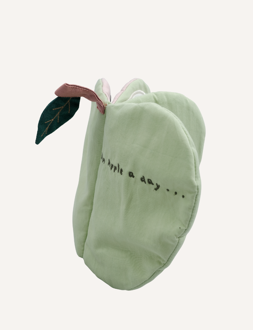 The Fabric Book - Green Apple by Fabelab is a soft, green apple-shaped fabric object crafted from organic cotton. It features a white flower and a small bee embroidered at its center, along with a tiny green leaf that has stitching details attached to the top, accompanied by a pink fabric loop. At the bottom, an off-white tag with text is visible—perfect for sensory play.