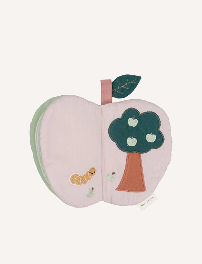 A child's hands touch the Fabric Book - Green Apple by Fabelab, an eco-friendly book designed like a sliced apple and made from organic cotton. The book features a green outer layer and a white inside with stitched details, including a worm and seeds. The child wears a light long-sleeve shirt, engaging in sensory play on a pale pink surface.