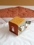 A soft fabric cube toy from Fabelab, featuring a hedgehog illustration on one side, a red and white mushroom on another, and a grey and orange fabric tag protruding from the top. The cube showcases a white, brown, and mustard color scheme with a label reading 