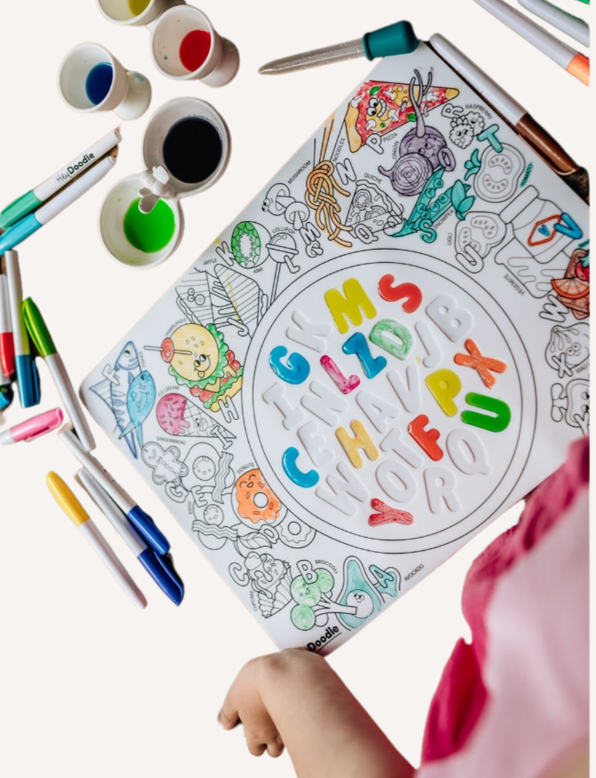 The image shows a HeyDoodle Sensory Mat - Alphabet Soup for children. The packaging displays outlined letters of the alphabet, and crayons are included to help develop fine motor skills. The background features a coloring scene with various images and characters.