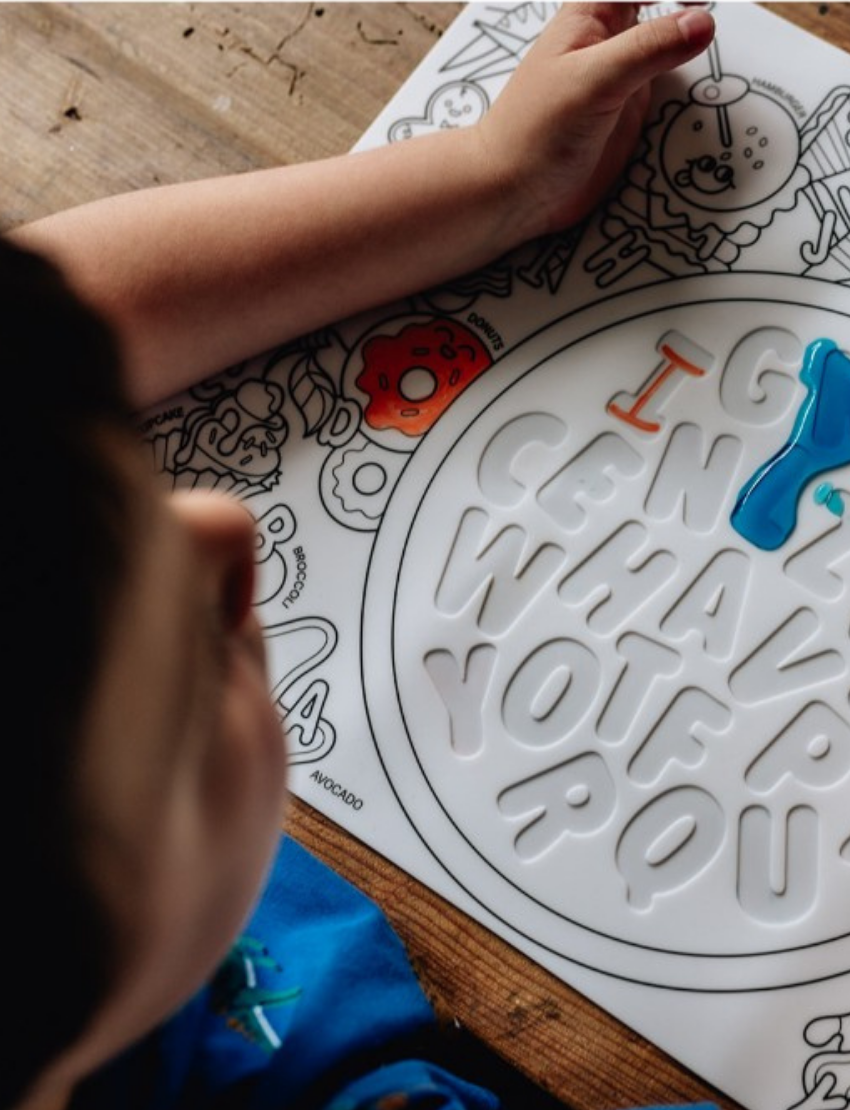 The image shows a HeyDoodle Sensory Mat - Alphabet Soup for children. The packaging displays outlined letters of the alphabet, and crayons are included to help develop fine motor skills. The background features a coloring scene with various images and characters.