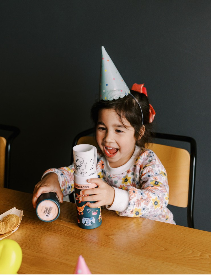 A cylindrical HeyDoodle container labeled "123 - Sugar & Spice" stands next to a partially colored-in silicone mat. The HeyDoodle Mini Mat, featuring illustrations of a unicorn, castle, fairy, mermaid, and various animals, shows some drawings colored while others remain in black and white.