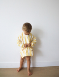 A toddler with short blond hair looks down at a Rommer Co Smock Bib in light blue and white stripes that they are holding. The child is wearing a shirt with matching light blue and white striped sleeves. The background is plain white.