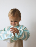 A toddler with short blond hair looks down at a Rommer Co Smock Bib in light blue and white stripes that they are holding. The child is wearing a shirt with matching light blue and white striped sleeves. The background is plain white.