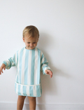 A toddler with short blond hair looks down at a Rommer Co Smock Bib in light blue and white stripes that they are holding. The child is wearing a shirt with matching light blue and white striped sleeves. The background is plain white.