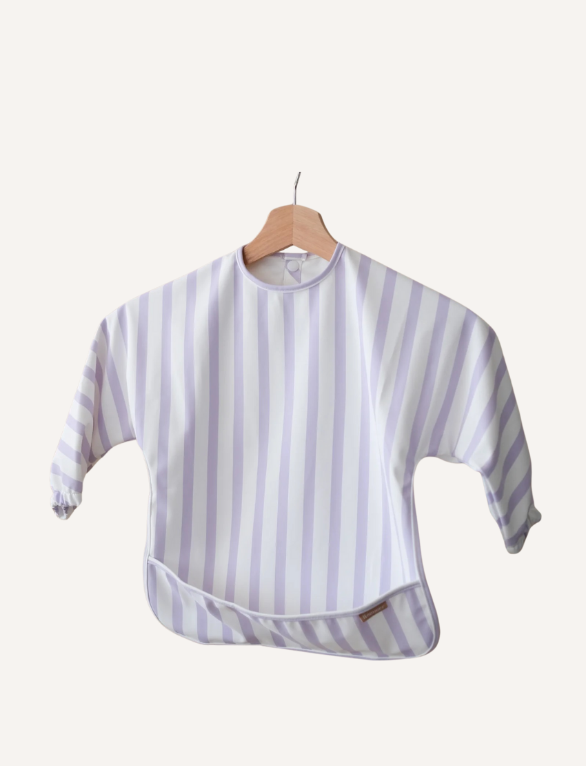 A toddler with short blond hair looks down at a Rommer Co Smock Bib in light blue and white stripes that they are holding. The child is wearing a shirt with matching light blue and white striped sleeves. The background is plain white.