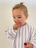 A toddler with short blond hair looks down at a Rommer Co Smock Bib in light blue and white stripes that they are holding. The child is wearing a shirt with matching light blue and white striped sleeves. The background is plain white.