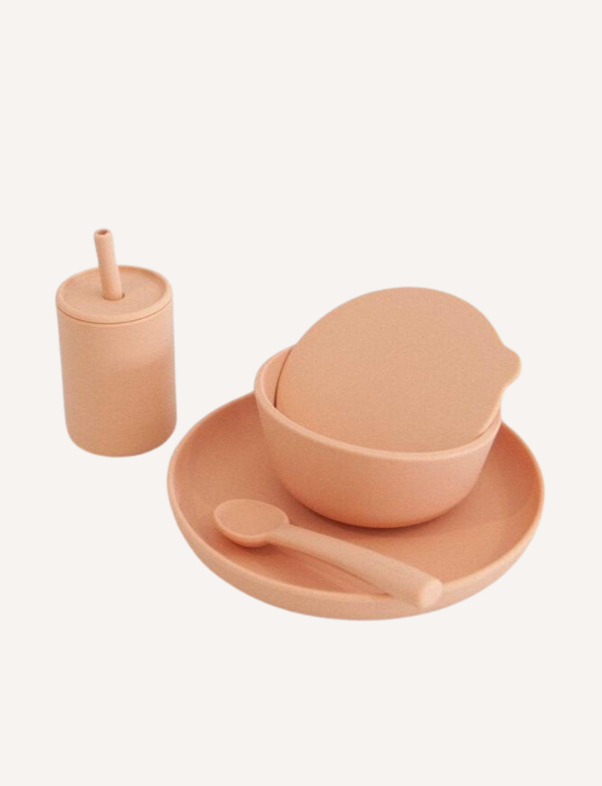 The Rommer Co Dinner Set is a peach-colored premium silicone set for children, featuring a cup with a straw, bowl with lid, plate, and spoon. Non-toxic and safe for the dishwasher and microwave, all items are neatly displayed against a plain white background.