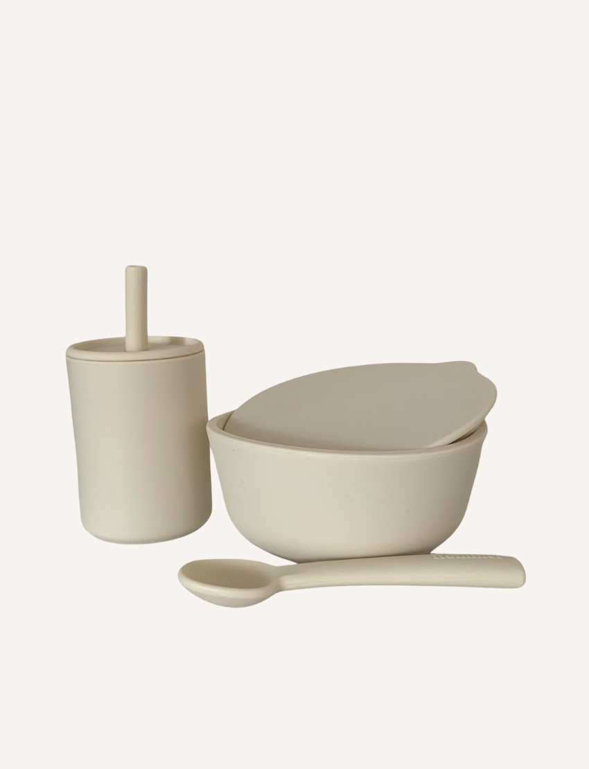 Rommer Co's Dinner Set is a minimalist collection of beige dishware, ideal as a baby gift. This set includes a lidded bowl, a cup with a straw, and a spoon. Crafted from premium silicone, the items are non-toxic and feature smooth surfaces and simple designs against an off-white background.